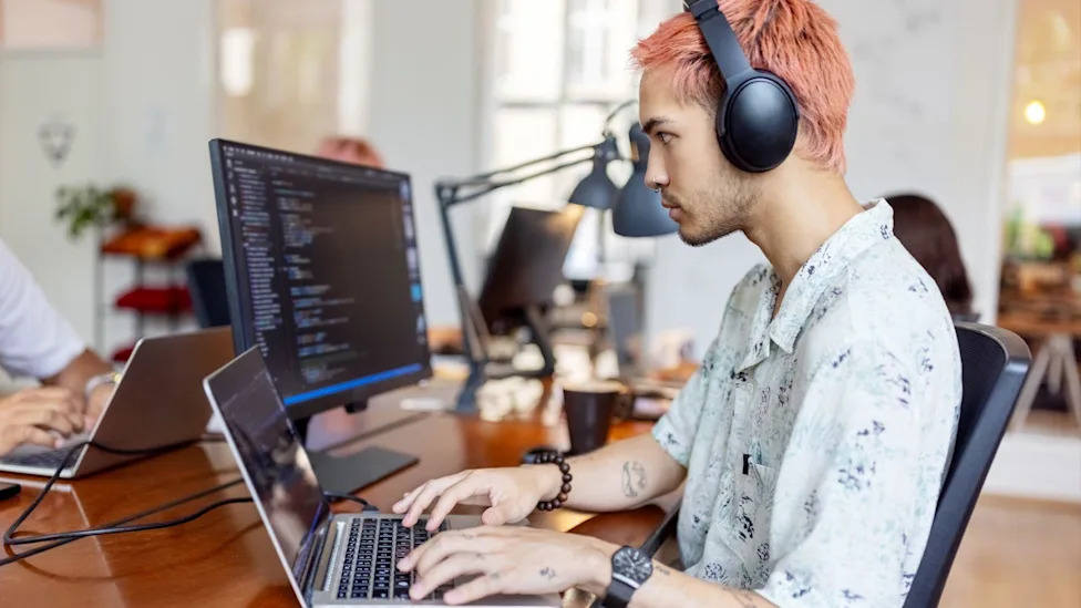 A tech worker with pink hair sits at a desk and works on a laptop