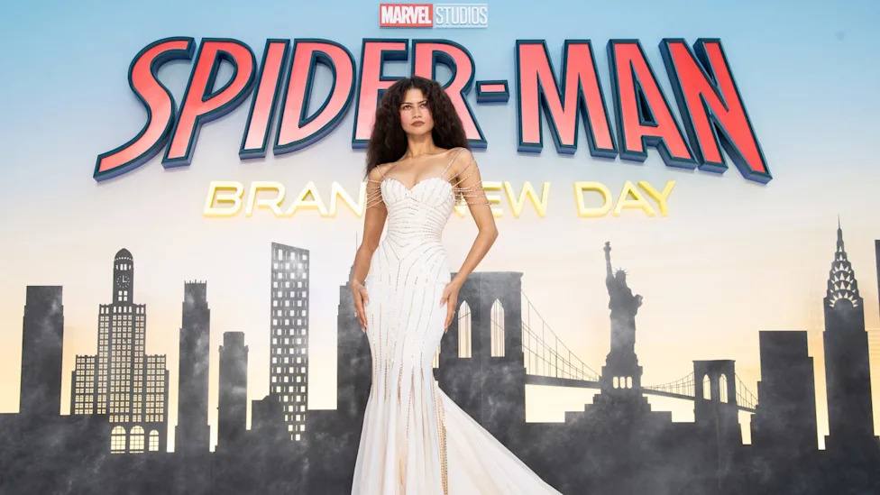 Zendaya in a glamorous white gown at a Spider-Man event, standing in front of a cityscape backdrop.