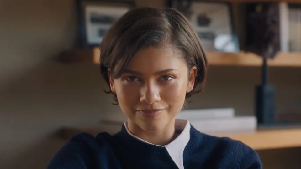Zendaya with short hair, wearing a navy blue sweater over a white shirt, smiling slightly, with shelves and framed photos in the background.