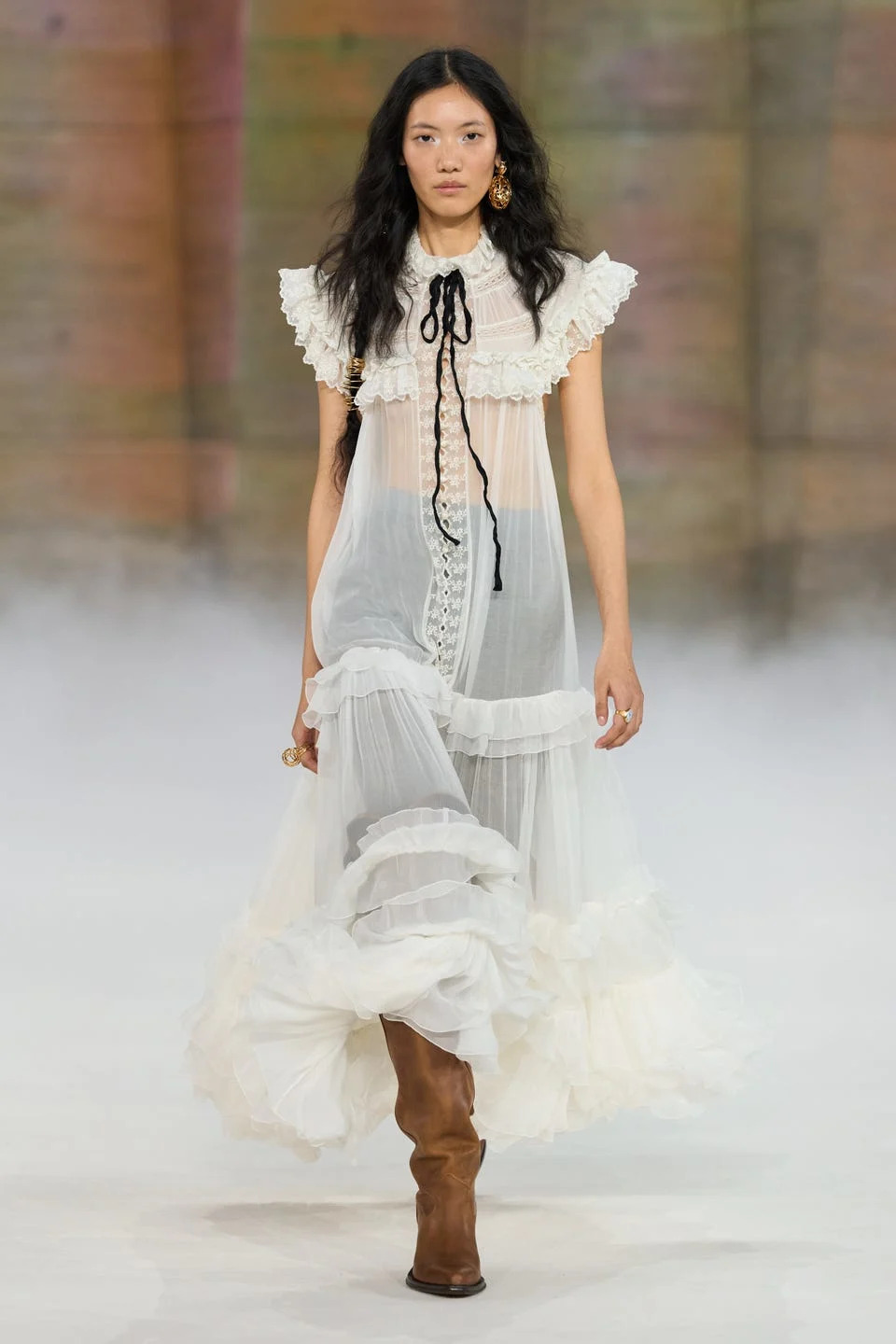Model walking in a flowing white dress with ruffles and brown knee-high boots on a runway background.