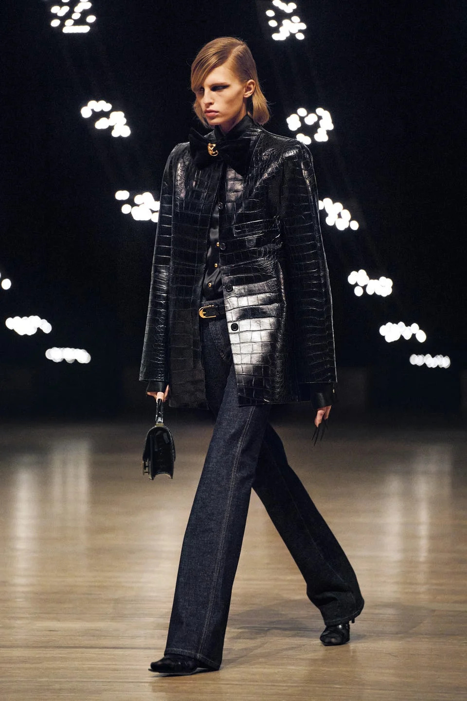 A model walks the runway wearing a black crocodile-print jacket and wide-leg jeans, with a small handbag in hand.
