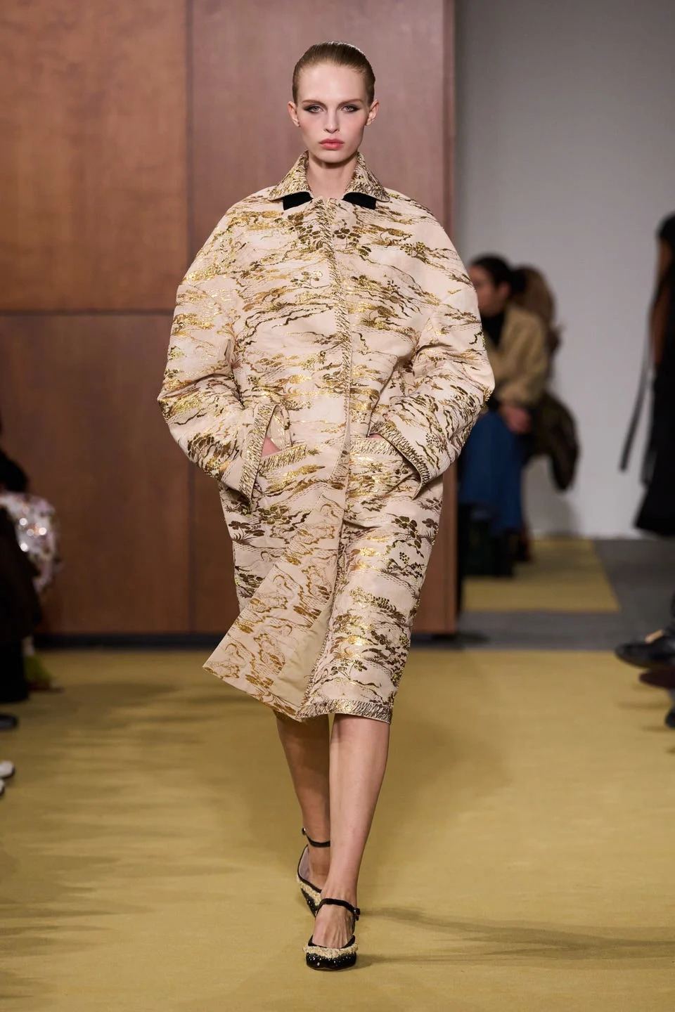 Model walking the runway in a patterned coat and stylish shoes, showcasing a contemporary fashion design.