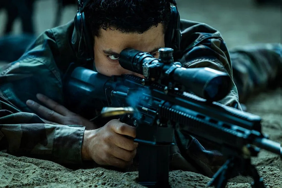 Person in camouflage gear and headphones aiming a rifle, lying on the ground.