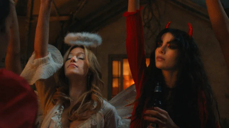 In a dimly lit house, a blonde woman is dressed in a white lace top with feathery wings and a fluffy halo headband with her eyes closed, and a woman with dark hair wearing a red top and red devil horns is holding a bottle of beer.
