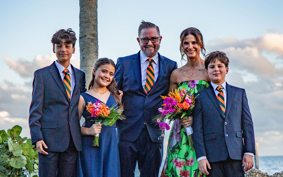 Outdoor group portrait of Ryan Goepel and family standing together near a waterfront location, with two holding floral bouquets and all dressed in formal attire.