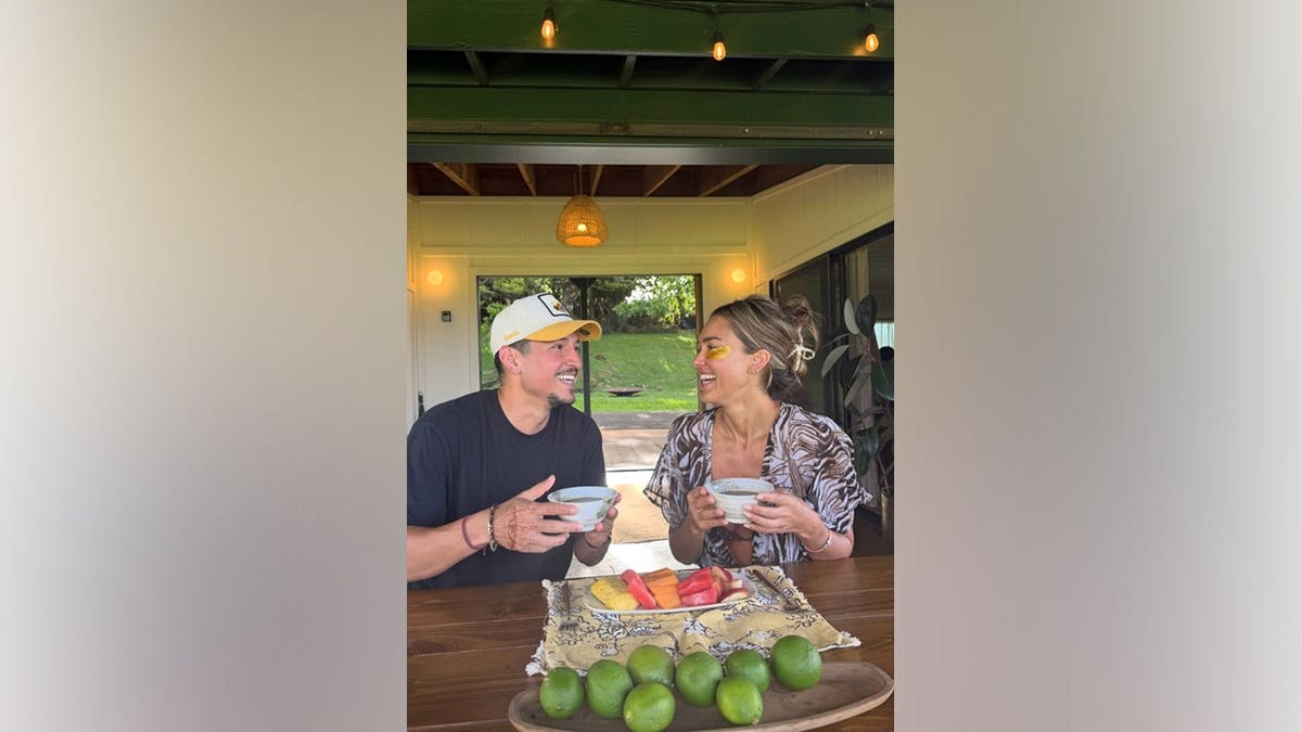 Jessica Alba and Danny Ramirez smile at each other while seated together at an outdoor table.