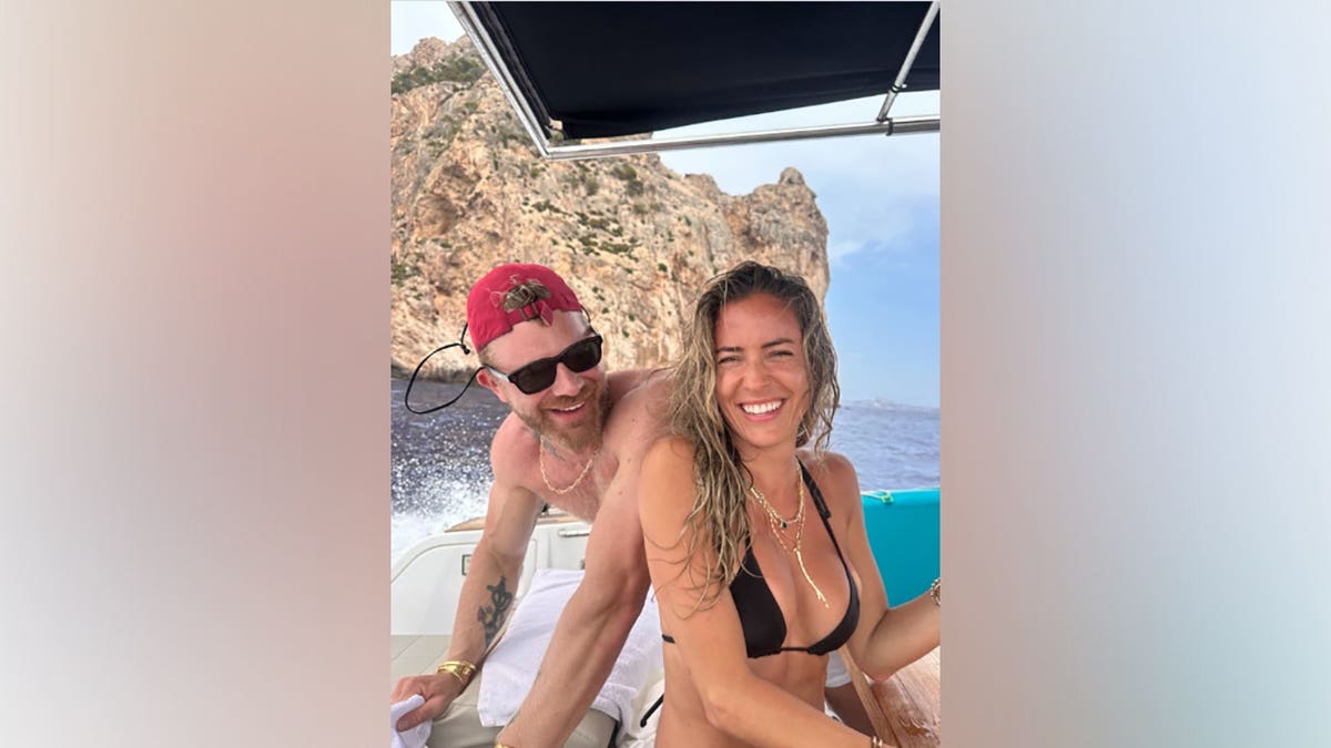 Kristin Cavallari, wearing a black bikini and sunglasses, smiles beside longtime friend Justin Anderson while aboard a boat surrounded by turquoise Mediterranean water.