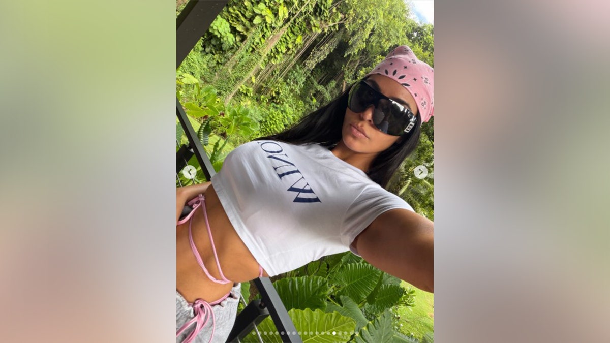 Kardashian posing in a white crop top with greenery behind her.