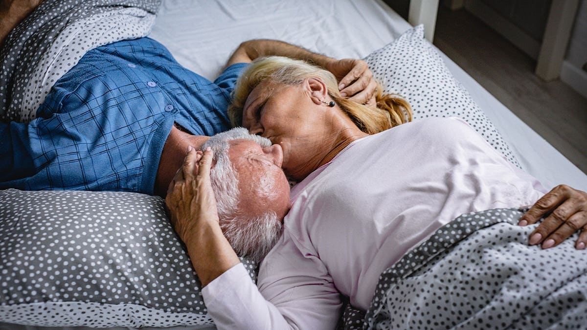 Senior man kisses senior woman while lying in bed together.