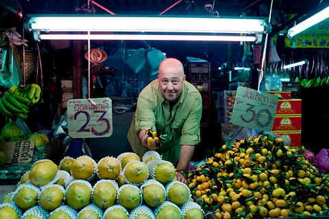 Courtesy of The Travel Channel Andrew Zimmern bends over a fruit stand and holds small orange fruits in his hand (Credit: Courtesy of The Travel Channel)