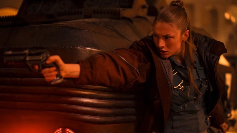 A woman with long blond hair tied partly in a bun is wearing a red/brown jacket, with some kid of harness on underneath it. She's crouching behind a futuristic car and is pointing a black pistol.