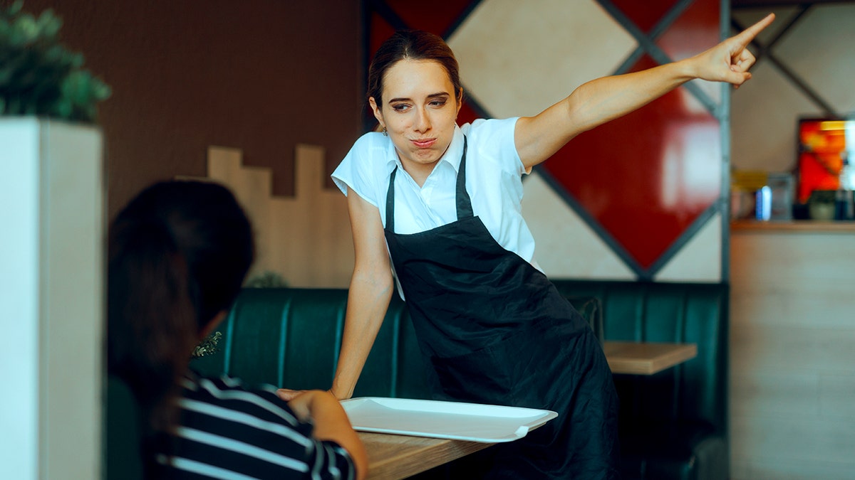 A waitress points toward the exit while speaking with a customer seated in a booth at a diner.
