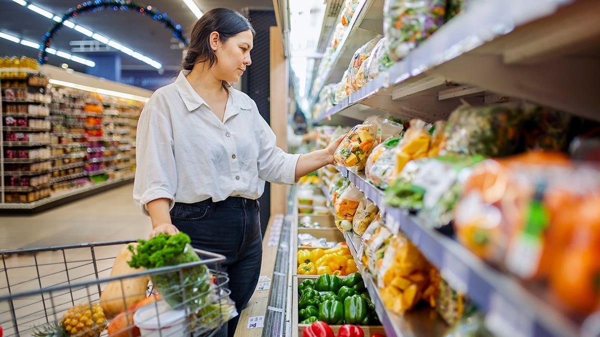 Woman shops for fresh vegetables in a supermarket produce aisle with a shopping cart.