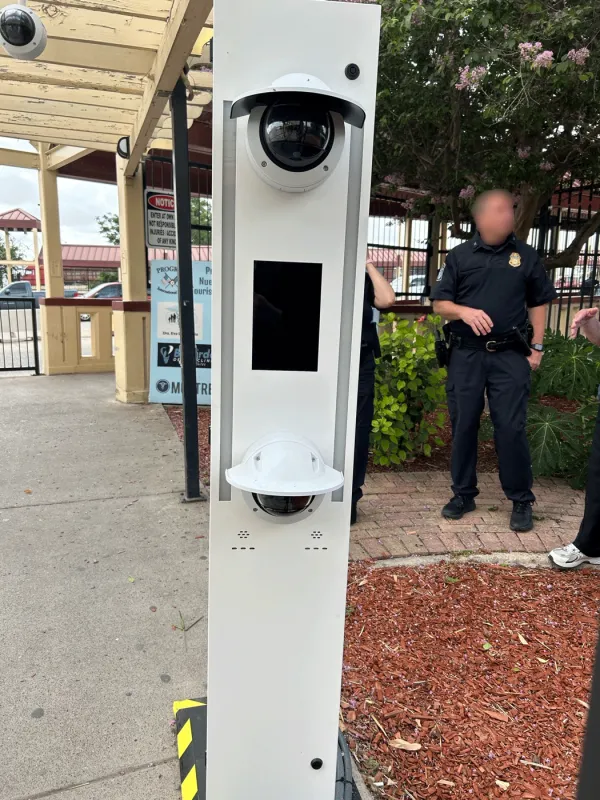 A close up of one of the upright grey metal kiosks. The upper and lower camera lenses are black and have a grey lid over them. In between the upper and lower cameras is a black rectangle that looks like it might be a screen of some sort. In the background to the right, a Border Patrol officer in a black uniform is talking with other individuals.