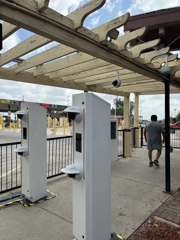 Two upright grey metal kiosks stand a few feet apart on either side of the walkway. The kiosks have one camera that is around the average height of an adult and one camera that is positioned about two feet lower. In the background, a man has passed through the camera areas and is walking away.