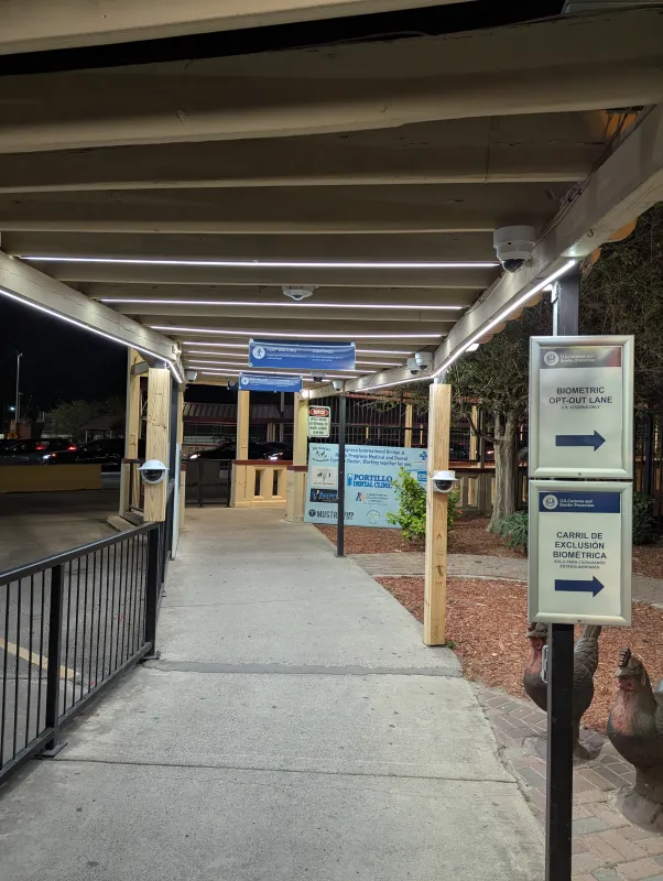 The walkway under the pergola at night with two signs indicating that there is a “Biometric opt-out lane” for U.S. Citizens Only. Looking down the walkway, we see the pergola is illuminated with lighting strips and has cameras in the rafters and on the poles.
