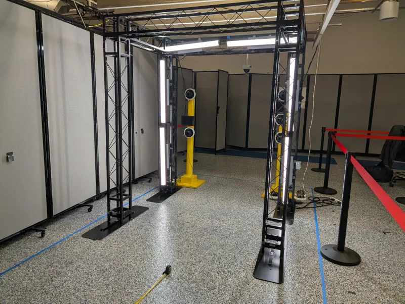 Inside at the MdTF, a prototype lighting structure set up for testing in a room. Black metal scaffolding creates a boxy entranceway with four stands supporting a metal square above for individuals to walk through. It stands about eight feet tall and has lights on it. There are yellow metal stands with cameras mounted on them on that far side of the structure.